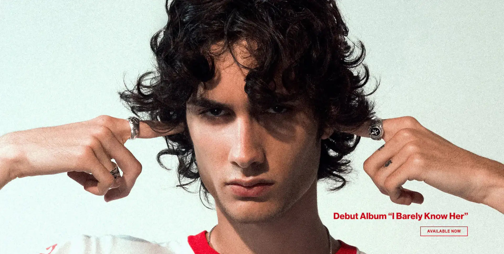 A young man with dark curly hair wearing a red and white shirt, adorned with silver rings on his fingers.